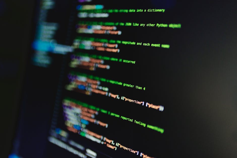 A person working at a desk with dual computer monitors, one displaying lines of code in a programming environment. The workspace is well-lit with natural light coming through large windows, showing an