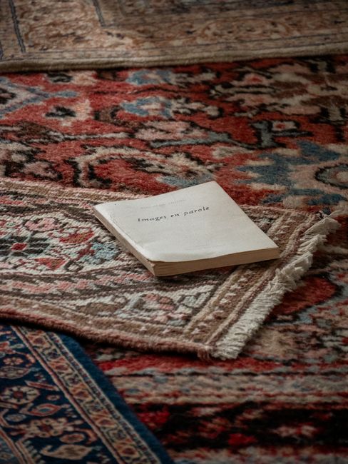Close-up of a professional vacuum cleaner with a transparent dust container filled with debris, connected to a flexible hose. The vacuum is positioned on a pink carpeted floor next to a wall with wood
